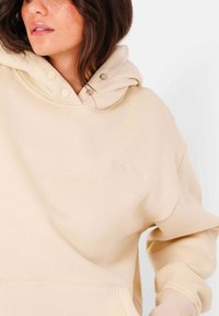 Felpa beige con cappuccio, caratterizzata da bottoni a pressione, una texture morbida e il nome del marchio ricamato sul davanti. Design oversize.