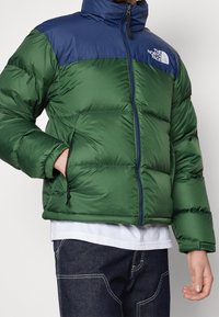 Groen en marineblauw gewatteerd jack met een hoge kraag, ritssluiting en elastische manchetten. Heeft zijzakken en een logo op de linkerborst.