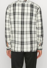 Rutig skjorta i vit, svart och grå flanell. Har en knappad krage och knäppta manschetter med springor på sidorna för ventilation.