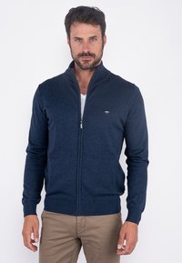 Cárdigan azul marino con cremallera hecho de tejido de punto suave; presenta una forma ajustada, puños acanalados y un pequeño emblema en el pecho.