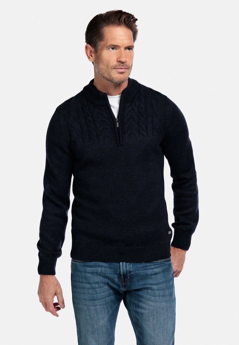 Man in een donkerblauwe kabelgebreide half-rits trui over een wit shirt en blauwe jeans, staande tegen een witte achtergrond.