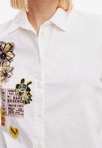 Bílá košile na zapínání s barevnou květinovou výšivkou a nášivkami, včetně motivu včely, s hladkou bavlněnou texturou.