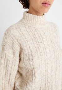 Beige Kabelstrickpullover mit hohem Kragen, der strukturierte Muster, kontrastierende Rippelemente und ein weiches, kuscheliges Material aufweist.