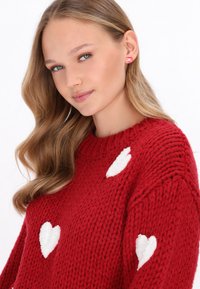 Roter gestrickter Pullover mit weißen Herzmustern, runder Ausschnitt und strukturierter Gestaltung. Getragen mit rosa Ohrsteckern und langen, welligen Haaren.