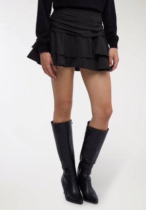 Minigonna nera a balze realizzata in tessuto liscio, con strati arricciati. Abbinata a stivali neri in pelle al ginocchio con punta appuntita.