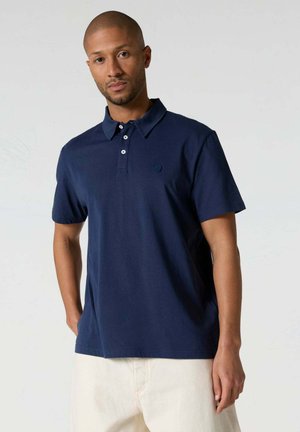 Hombre con el pelo corto que lleva una camisa polo de manga corta azul marino y pantalones color blanco roto, de pie con una mano en el bolsillo.