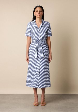 Vrouw staand in een midi-jurk met diagonale blauw-witte strepen, korte mouwen en een ceintuur die is gestrikt, met gouden sandalen met bandjes.