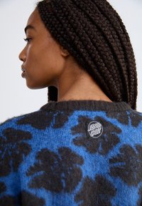 Maglione in maglia nero e blu, con un motivo floreale e un'etichetta con il marchio sul retro. La texture appare morbida e pelosa.