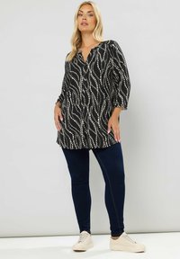 Svart tunika med vitt vågmönster, knäppning fram, och ¾ ärmar, parat med mörkblå tajta jeans och beige sneakers.