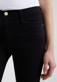 Jeans de mezclilla negros con un diseño ajustado, que presentan un botón dorado en la cintura y detalles de costura sutiles en los bolsillos. Textura suave.