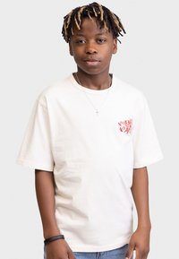 T-shirt en coton blanc à manches courtes, affichant le texte rouge "NO HEART NO A" sur le côté gauche. Associé à un jeans bleu et à un bracelet noir.