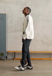 Weißer, fuzzy Pullover mit schwarzen Streifen, schwarze Sporthose und schwarze Turnschuhe mit weißen Akzenten, stehend neben einem grauen Spind.