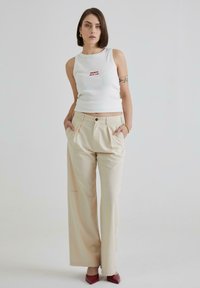 Hvid ribbet tanktop med rød tekst, parret med højhalsede beige brede bukser; modellen bærer spidse røde hæle og har kort hår.