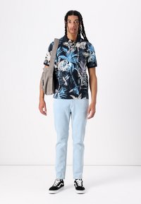Un polo bleu marine à manches courtes avec des motifs de feuilles tropicaux bleus et blancs, associé à un jean bleu clair et des baskets noires à tige basse.