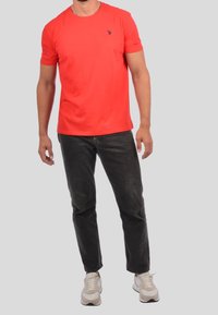 Punainen puuvillapaita, jossa on pyöreä kaula-aukko, lyhyet hihat ja pieni tummansininen logo vasemmassa rinnassa. Yhdistetty tummaan farkkuun ja tennareihin.