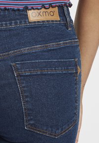 Jeans de mezclilla azul oscuro con una cintura alta, costuras naranjas y un parche de cuero con marca. Presenta un diseño de bolsillo trasero.