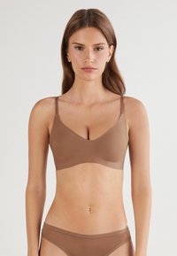 Vrouw met lang bruin haar in een bijpassende naadloze bruine bh en ondergoed tegen een effen lichte achtergrond.