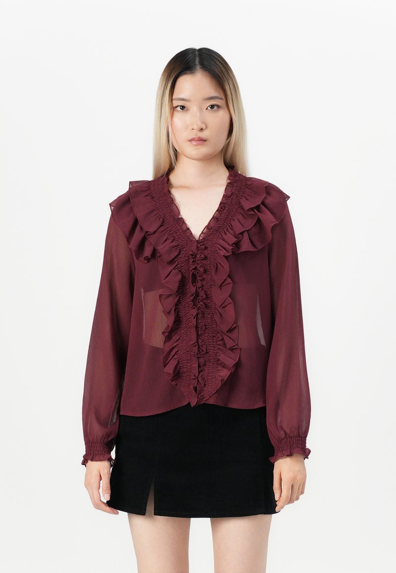 Blusa trasparente bordeaux con scollatura a volant e pattina frontale, maniche lunghe con polsini elastici, abbinata a una gonna nera corta.