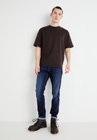 Brown cotton t-shirt, relaxed fit, short sleeves; paired with dark blue slim-fit jeans and gray lace-up boots with yellow accents.