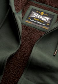 Groene zip-up hoodie met een bruine fleece binnenkant. Het label leest "Superdry Supply Co." met gouden accenten. Gelaagd stof en bronzen rits.