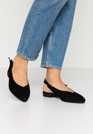Person iført lyseblå jeans og sorte slingback-flats med lav hæl og spidse tæer på et hvidt gulv.