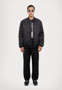 Fekete steppelt dzseki álló gallérral, grafikai mintájú pólóval és fekete nadrággal párosítva. A modellen fehér téglalap alakú napszemüveg és fekete sportcipő van.