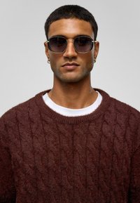 Ronde zonnebrillen met donkere lenzen, gouden metalen frame. Model draagt een bordeauxrode kabeltrui over een witte t-shirt. Kort donker haar.
