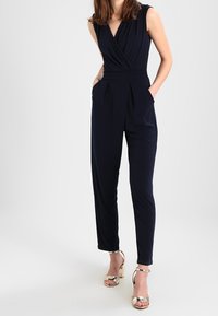 Combinaison bleu marine à encolure en V, sans manches, avec un devant plissé. Associée à des sandales à talons hauts métalliques dorées, mettant en valeur l'ensemble.