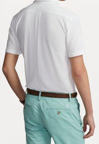 Polo shirt bianco a maniche corte, texture liscia, indossato con pantaloni turchesi chiari, caratterizzati da una cintura marrone e un branding sobrio sui pantaloni.