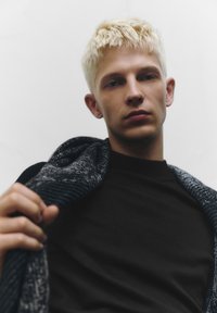 Svart stickad tröja med en texturerad, grå halsduk hängande över axeln. Modellen har kort, blekt blont hår och ett neutralt uttryck.