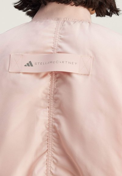 Pink bomber jacket features a smooth texture, elastic collar, and a prominent stitched logo label on the back. Minimalist design.
