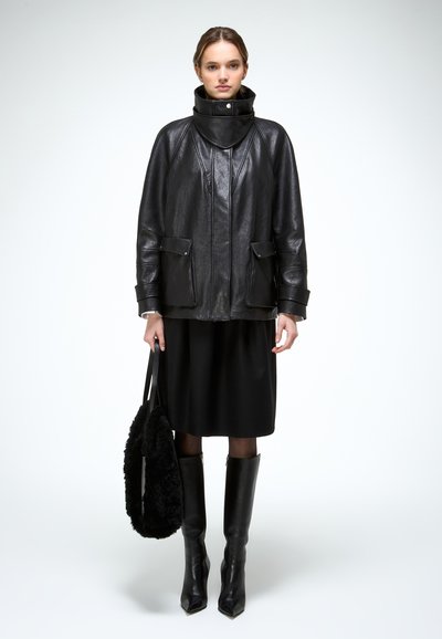 Mujer con chaqueta de cuero negra de cuello alto, falda negra, botas de cuero hasta la rodilla y bolso de piel negro, posando contra un fondo liso.