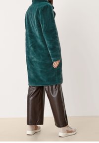 Turkusowy płaszcz z sztucznego futra z wysokim kołnierzem, prostym krojem i długimi rękawami. Połączony z brązowymi spodniami z szerokimi nogawkami z sztucznej skóry i beżowymi trampkami.