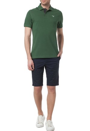 Polo vert à col court, assorti avec un short bleu marine. La tenue est complétée par des chaussures de sport blanches. Tissu et coupe décontractés.