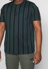 Hombre que lleva una camiseta negra con rayas verticales verdes y azules y pantalones cargo verde olivo con bolsillo con cremallera.
