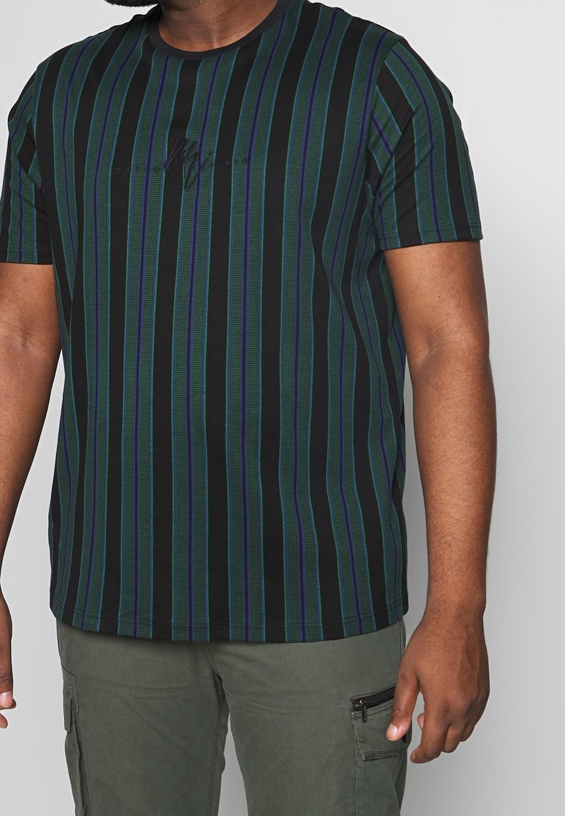 Hombre que lleva una camiseta negra con rayas verticales verdes y azules y pantalones cargo verde olivo con bolsillo con cremallera.
