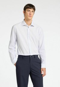 Camicia bianca a righe con colletto classico e maniche lunghe, abbinata a pantaloni scuri. Presenta una texture di tessuto liscia.