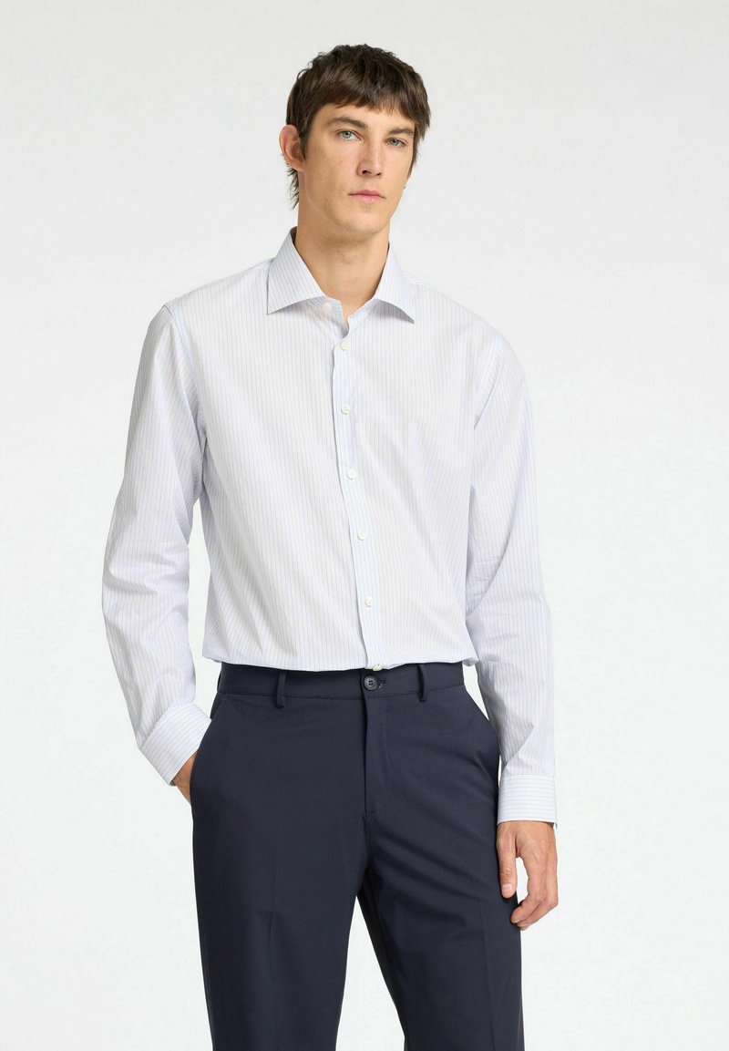 Camicia bianca a righe con colletto classico e maniche lunghe, abbinata a pantaloni scuri. Presenta una texture di tessuto liscia.