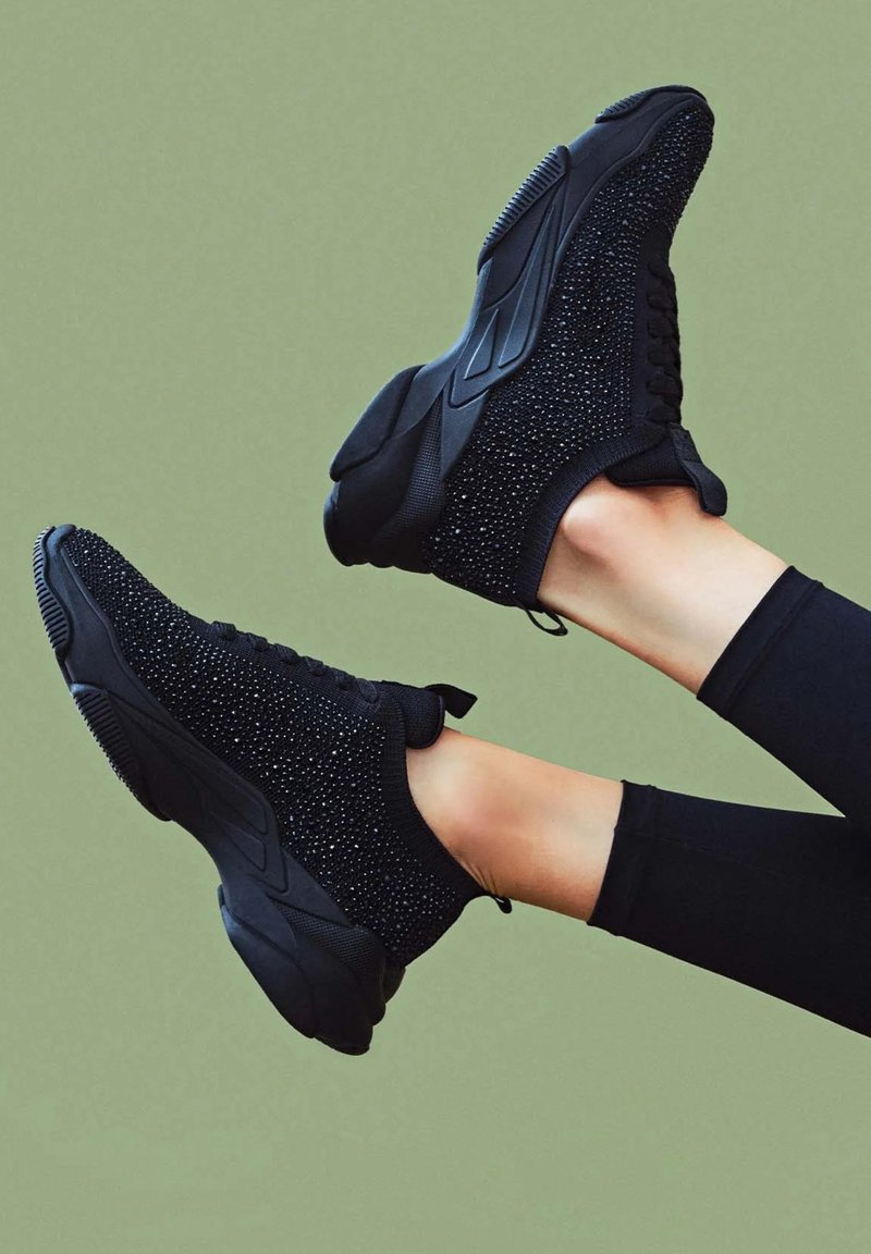 Black sneakers with a textured upper adorned with small studs, featuring a flexible sole and a slip-on design.