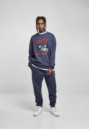 Mann mit geflochtenem Haar, der einen marineblauen Starter-Sweatshirt und passende Jogginghose sowie weiße Turnschuhe trägt und vor einem einfarbigen Hintergrund steht.