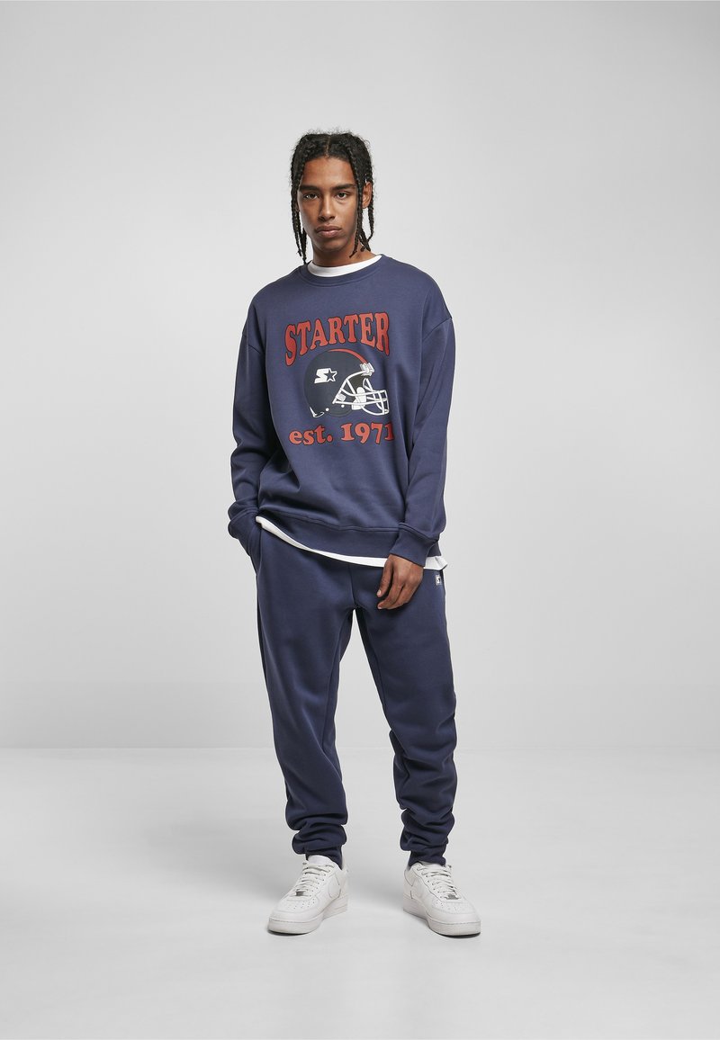 Vīrietis ar koptām bizēm, tērpies tumši zilā Starter džemperī un atbilstošos sporta svārkos, ar baltām sporta kurpēm, stāv pret vienkrāsainu fonu.