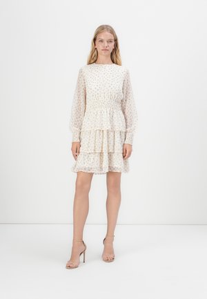 Floral-patterned, cream-colored dress with long sleeves, smocked waist, and tiered ruffle skirt. Paired with beige strappy heels.