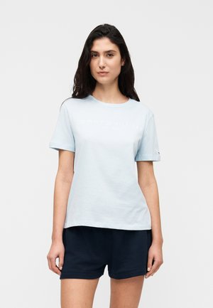 Donna con lunghi capelli scuri che indossa una maglietta azzurro chiaro a maniche corte e pantaloncini scuri, in piedi davanti a uno sfondo chiaro e uniforme.