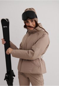 Beige skijakke med høj krave, lynlås og lommer. Parret med matchende bukser, holder sorte ski og bærer en strikket beige hue.
