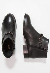 Botas de cuero negro hasta el tobillo con cremallera lateral y detalle decorativo de cadena de metal oscuro, una bota en posición vertical y la otra vista desde arriba.