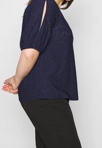 Námořnický modrý top s perforovaným květinovým vzorem, krátké nabírané rukávy s bočními výřezy, volný střih, vyrobený z lehkého materiálu.