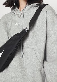 Osoba v šedé mikině Nike s stříbrným řetízkem a černou krosnou s bílým logem Nike.