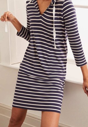 Kobieta ubrana w długi, granatowo-biały pasiasty sweter z kapturem i sznureczkami, siedząca obok białej wewnętrznej ramy okna.