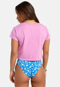 Donna con lunghi capelli castani che indossa una maglietta rosa corta e pantaloni bikini blu con fiori, voltata di spalle su uno sfondo neutro.