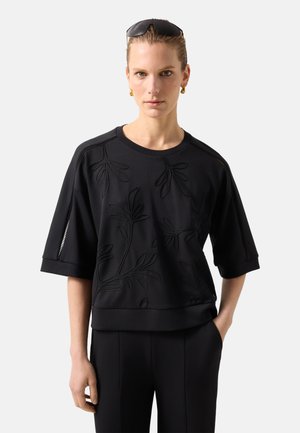 Kvinde iført sort kortærmet top med blomsterbroderi og sorte bukser, solbriller på hovedet og guld hoop øreringe.